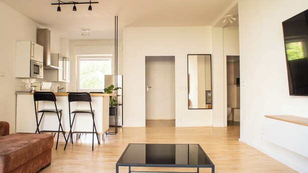 Una sala de estar moderna con una mesa de centro de vidrio, un sofá marrón y un comedor con dos sillas, con una cocina al fondo.