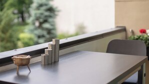 Una mesa con un cenicero de metal y objetos cilíndricos, una ventana con vistas a la vegetación, y una silla.