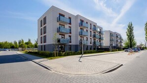 Un immeuble résidentiel moderne à plusieurs étages, doté de balcons, d’une allée pavée et surplombant un ciel dégagé.