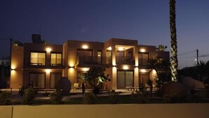A modern two-story house with a well-lit exterior, large windows, and a landscaped garden with palm trees and shrubs. - La Rêve Luxury Apartments (Hersonissos)
