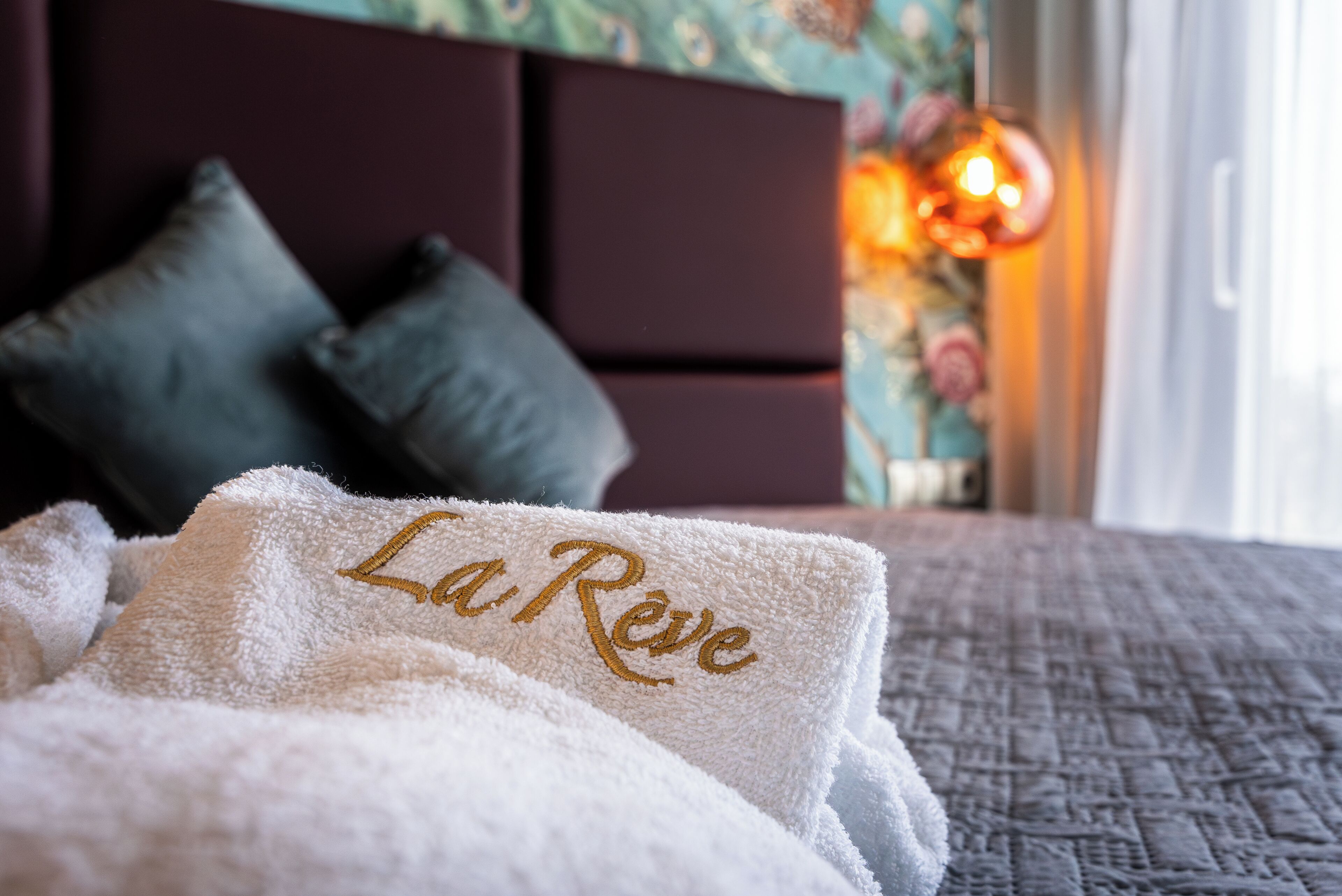 A neatly made bed with a white towel featuring embroidered text, a dark headboard, and a decorative wall with a floral pattern.