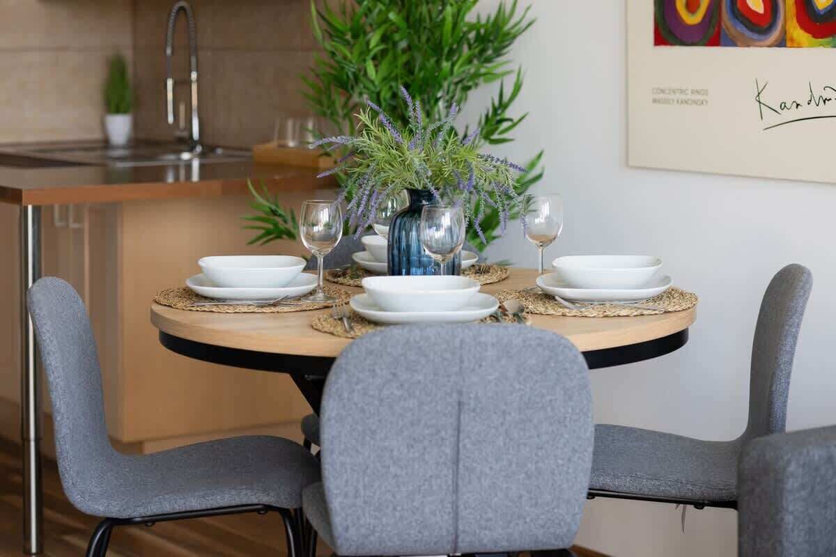 Un coin repas avec une table ronde dressée pour quatre personnes, entourée de chaises grises, et agrémenté d’un vase de verdure et de verres à vin posés sur la table.