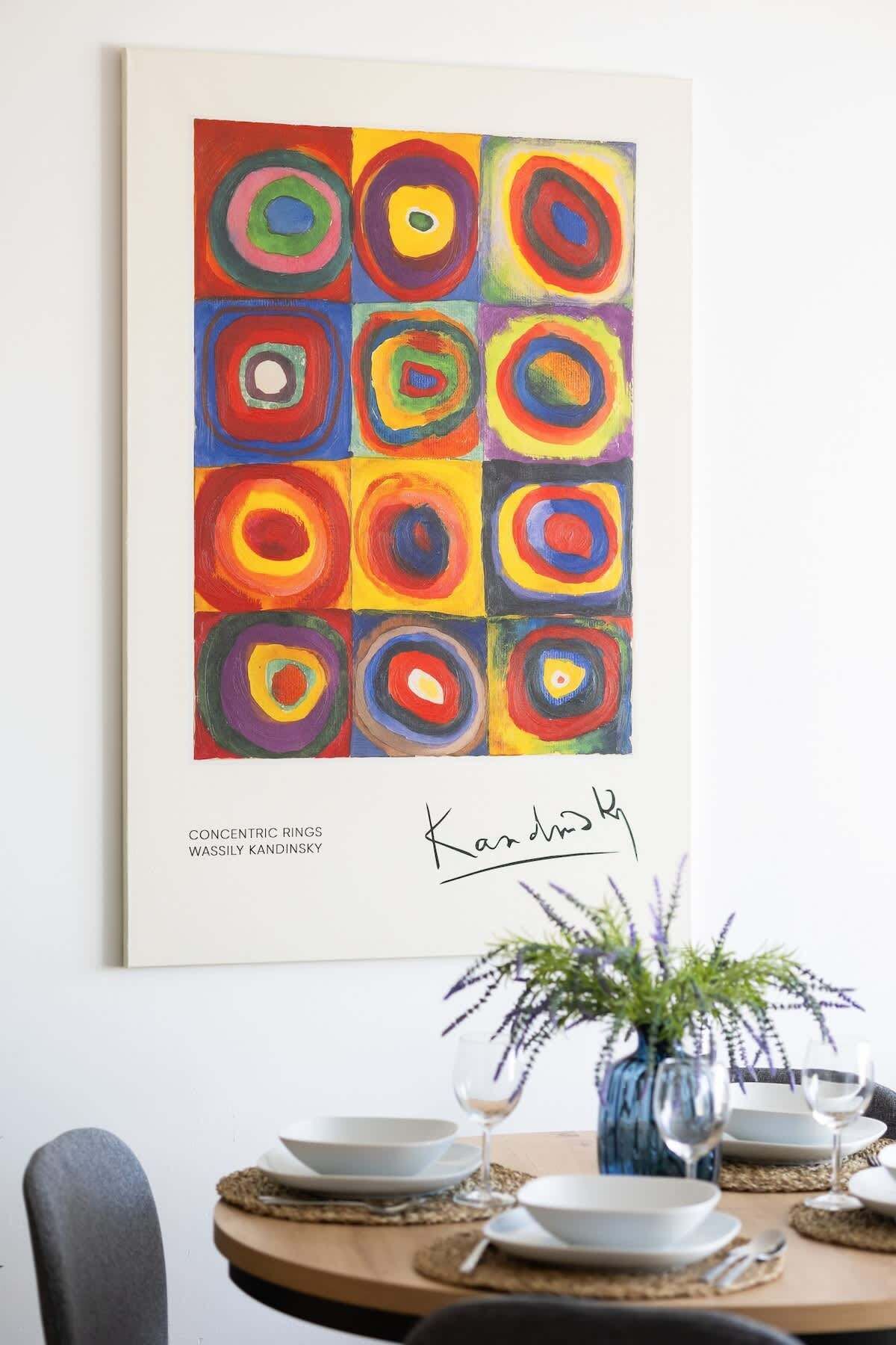 A framed artwork titled 'Concentric Rings' by Wassily Kandinsky, signed by the artist, displayed above a dining table with a vase of flowers and dinnerware.