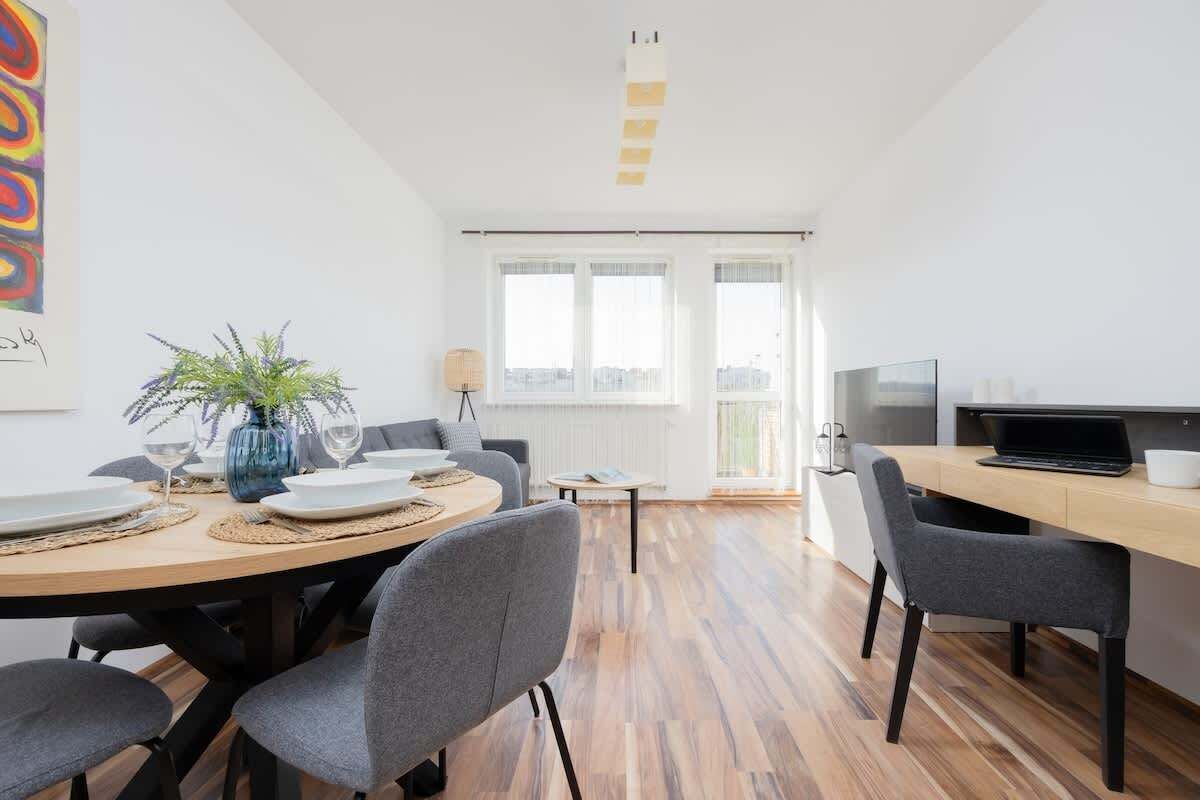 Una habitación moderna y luminosa con una mesa de comedor para cuatro personas, un ordenador portátil sobre un escritorio y un ventanal con vistas a la ciudad.
