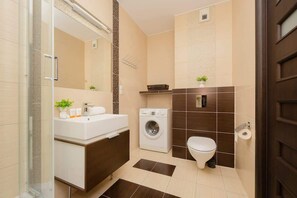 Een moderne badkamer met een douchecabine van glas, een wit wastafelmeubel met een donker aanrechtblad, een wasmachine en een toilet.