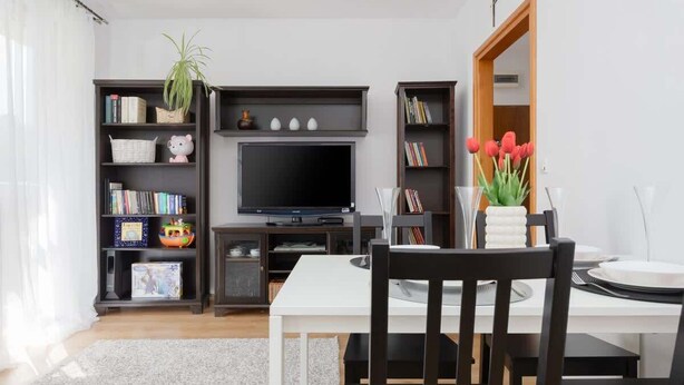 A modern living room with a flat-screen TV, a dining table with chairs, and built-in bookshelves.