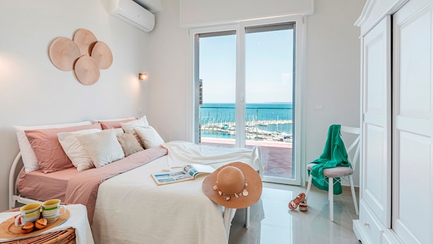 Un dormitorio con cama, ventana con vista a la playa, una silla y una mesa con un libro y un sombrero.