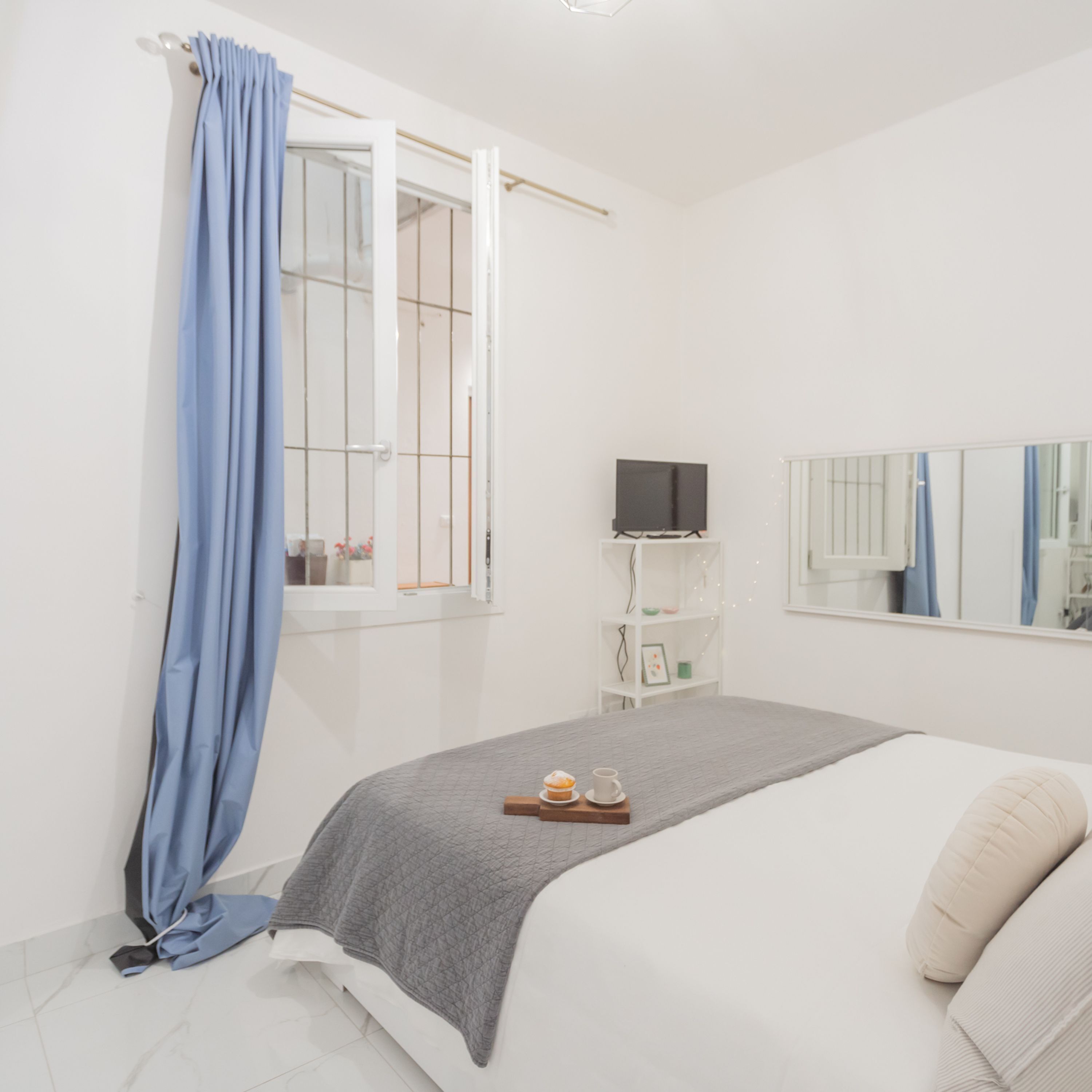 Een slaapkamer met een bed, een televisie, een raam met gordijnen, een nachtkastje en een spiegel.