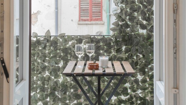 A table with two wine glasses and a candle, set against a backdrop of green foliage and a red shuttered window. - Luce Apartment in Como (Como)