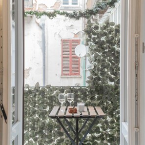 A table with two wine glasses and a candle, set against a backdrop of green foliage and a red shuttered window. - Luce Apartment in Como (Como)