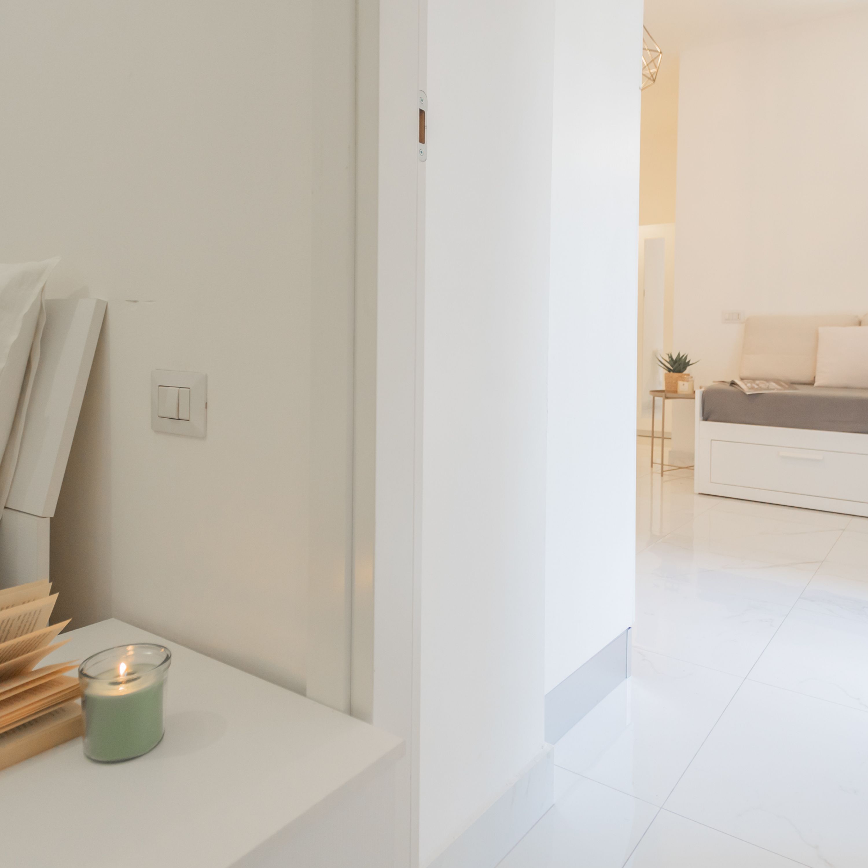 A modern, minimalist room with a white wall, a small table with a candle, and a glimpse of a bed in the background.