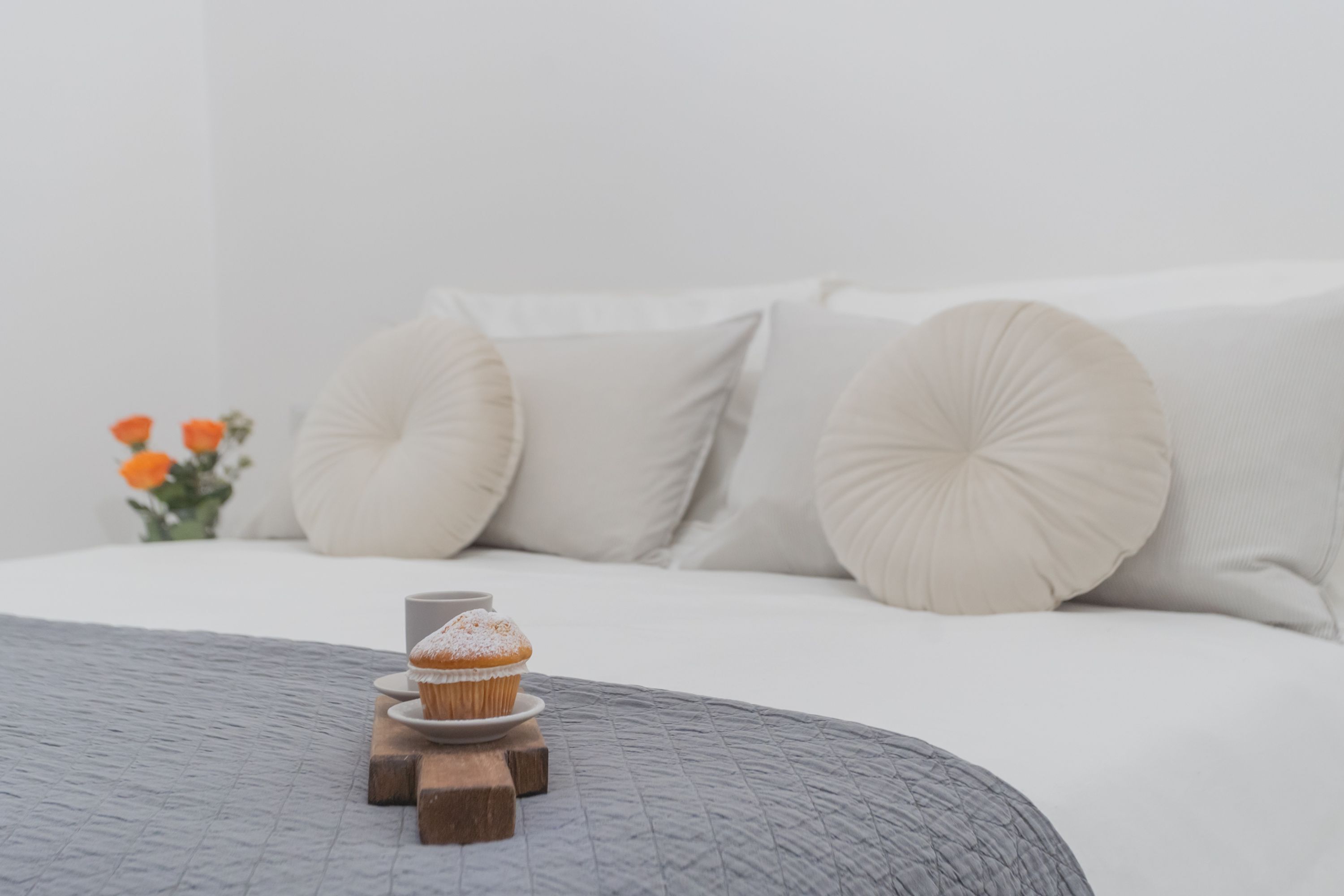 A neatly made bed with a grey quilt, a wooden stand holding a cupcake, and a vase with orange flowers on the side.