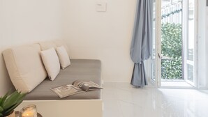 A modern, minimalist room with a grey sofa, a small side table with a candle and a plant, and a view of greenery through an open door. - Luce Apartment in Como (Como)
