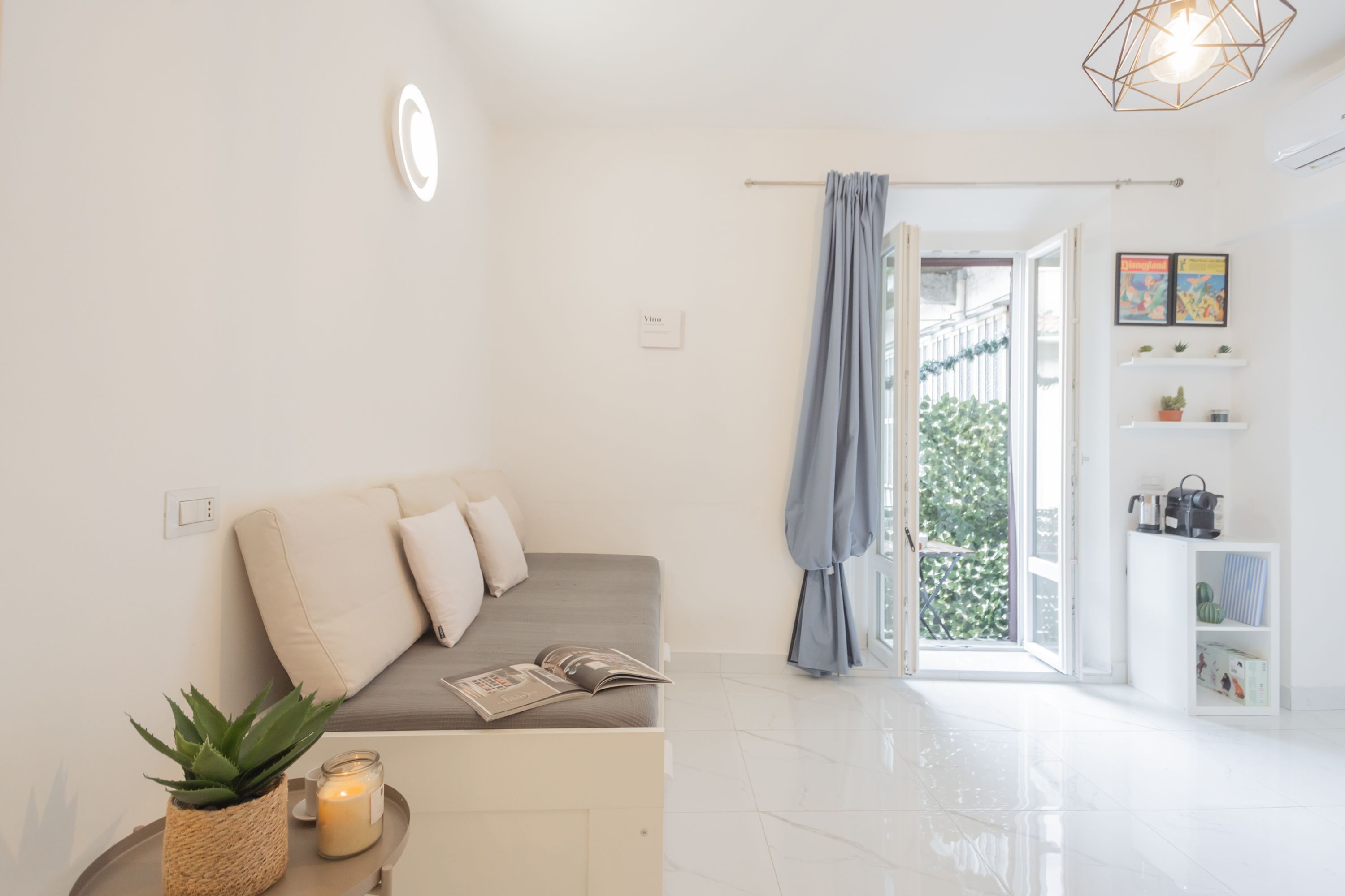 A modern, well-lit room with a sofa, a small table with a candle and a plant, and a view of the outdoors through an open door.