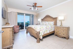 A hotel room with a bed, a dresser, a chair, a ceiling fan, and a view through a large window.