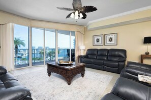 A spacious living room with a large window, a ceiling fan, a black leather sofa, a wooden coffee table, and a view of the city.