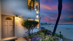 A coastal house with a well-lit entrance, a small garden with purple flowers, and a view of the beach and city skyline at dusk. - Bayfront Oasis Near Boardwalk and Water (San Diego)