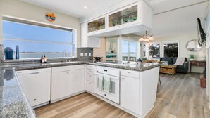 A modern kitchen with granite countertops, white cabinetry, and a large island. There is a window with a view of the water, and the kitchen connects to a living area with a sofa and a coffee table. - Bayfront Oasis Near Boardwalk and Water (San Diego)