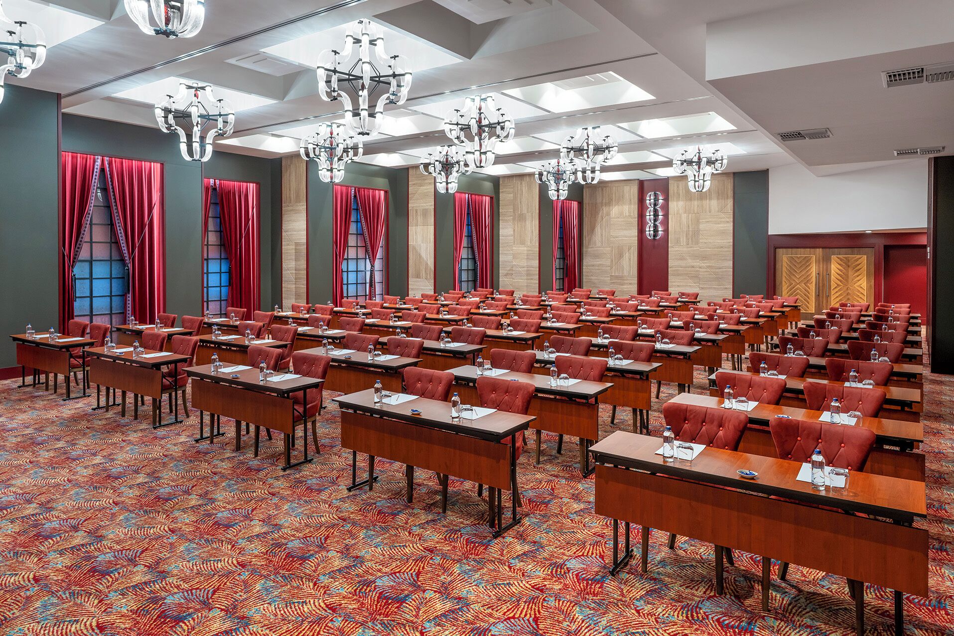 Un salón de conferencias con filas de mesas y sillas, ventanas amplias con cortinas rojas y una alfombra estampada.