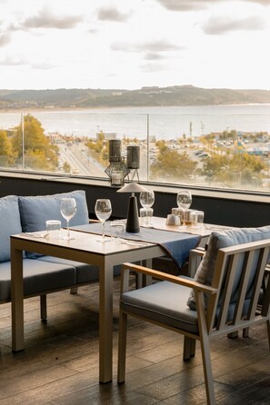 Un comedor con una mesa dispuesta para dos personas, vista al agua y un paisaje urbano de fondo.