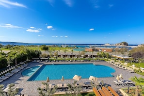 Zona de piscina en un resort con múltiples tumbonas y sombrillas, rodeada de vegetación y vistas panorámicas del mar y colinas distantes.
