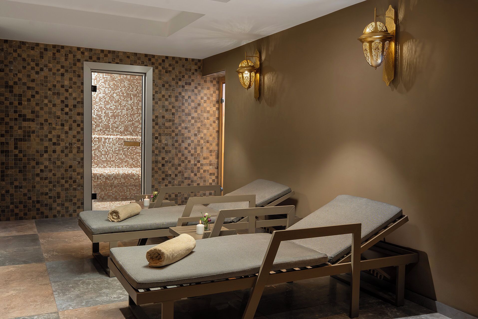 A spa room with three massage tables, a tiled wall, and two wall-mounted lamps.
