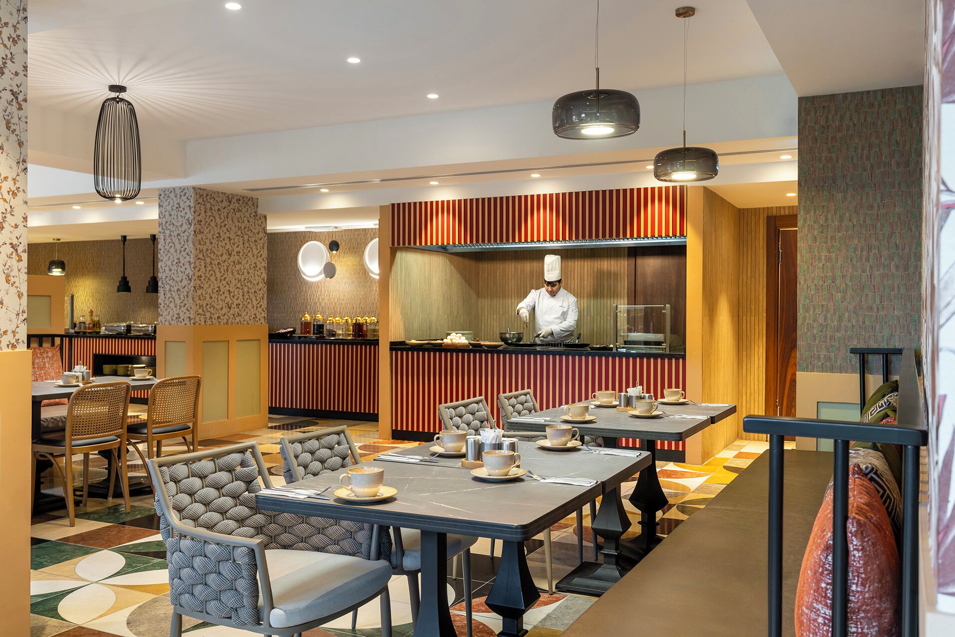 A restaurant with a chef preparing food, tables with chairs, and a bar area.