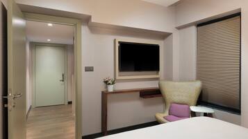 A hotel room with a bed, a desk with a monitor, a chair, and a window with blinds.
