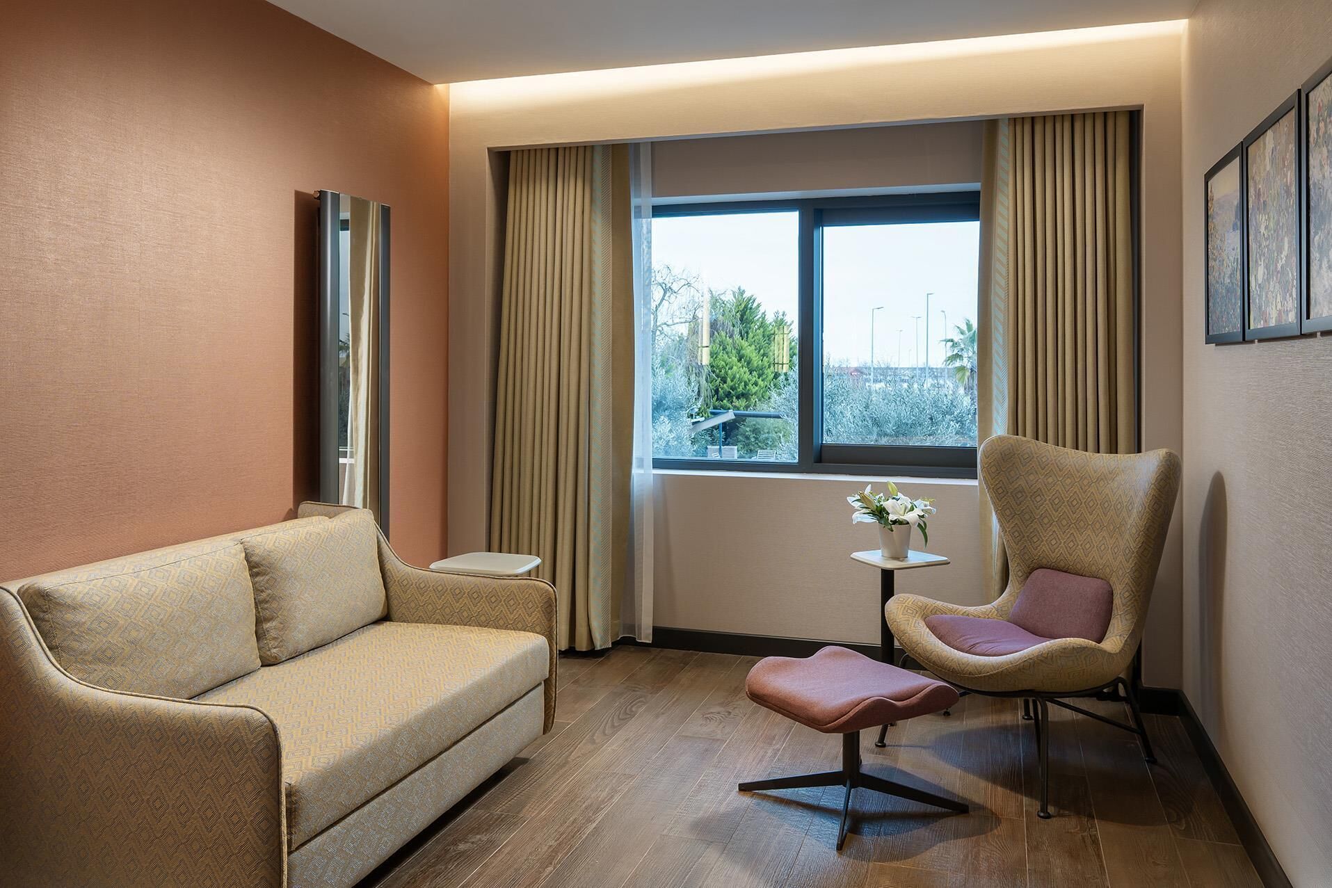 A hotel room with a sofa, a chair, a small table with a vase, and a window with curtains.