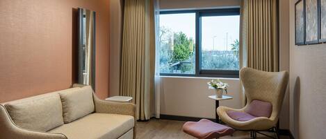 A hotel room with a sofa, a chair, a small table with a vase, and a window with curtains.