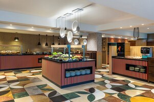 A modern hotel lobby with a central buffet area, a coffee station, and a seating area with a patterned floor.