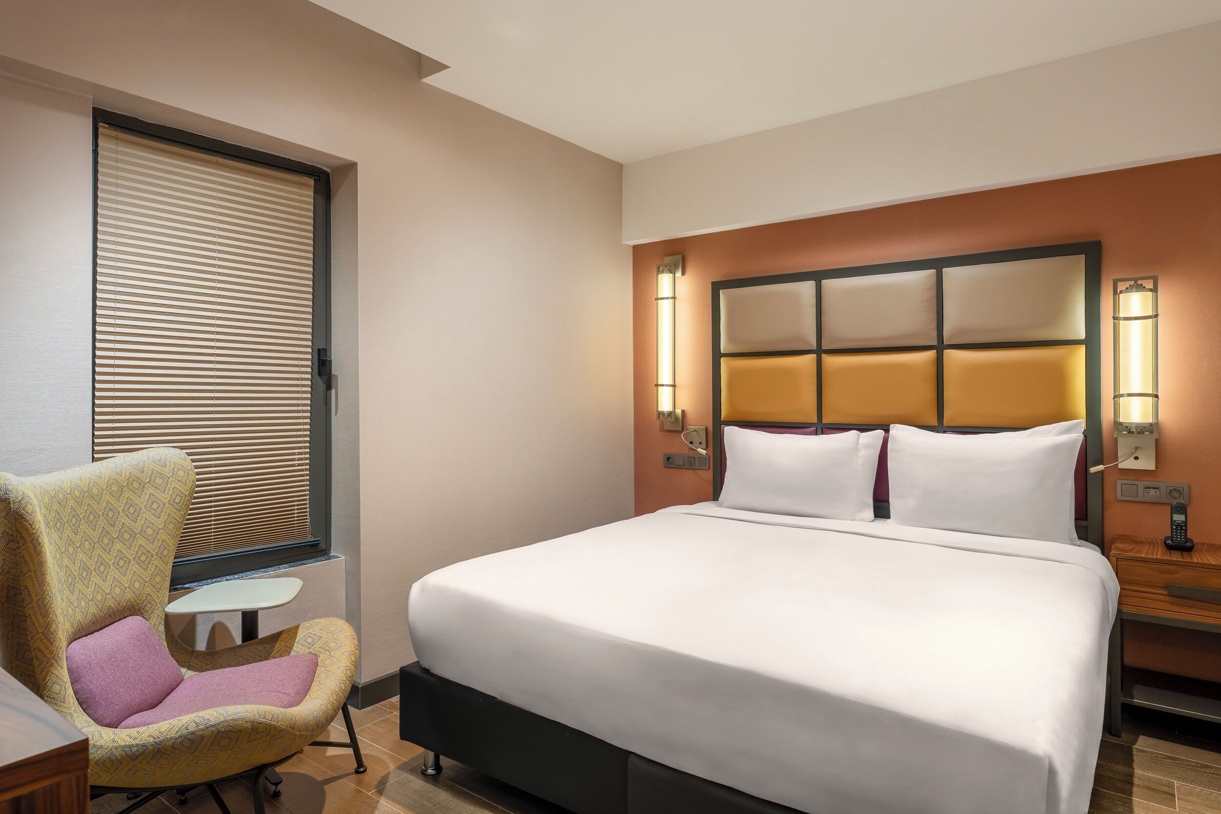A hotel room with a large bed, a chair, a small table, and two wall-mounted lamps.