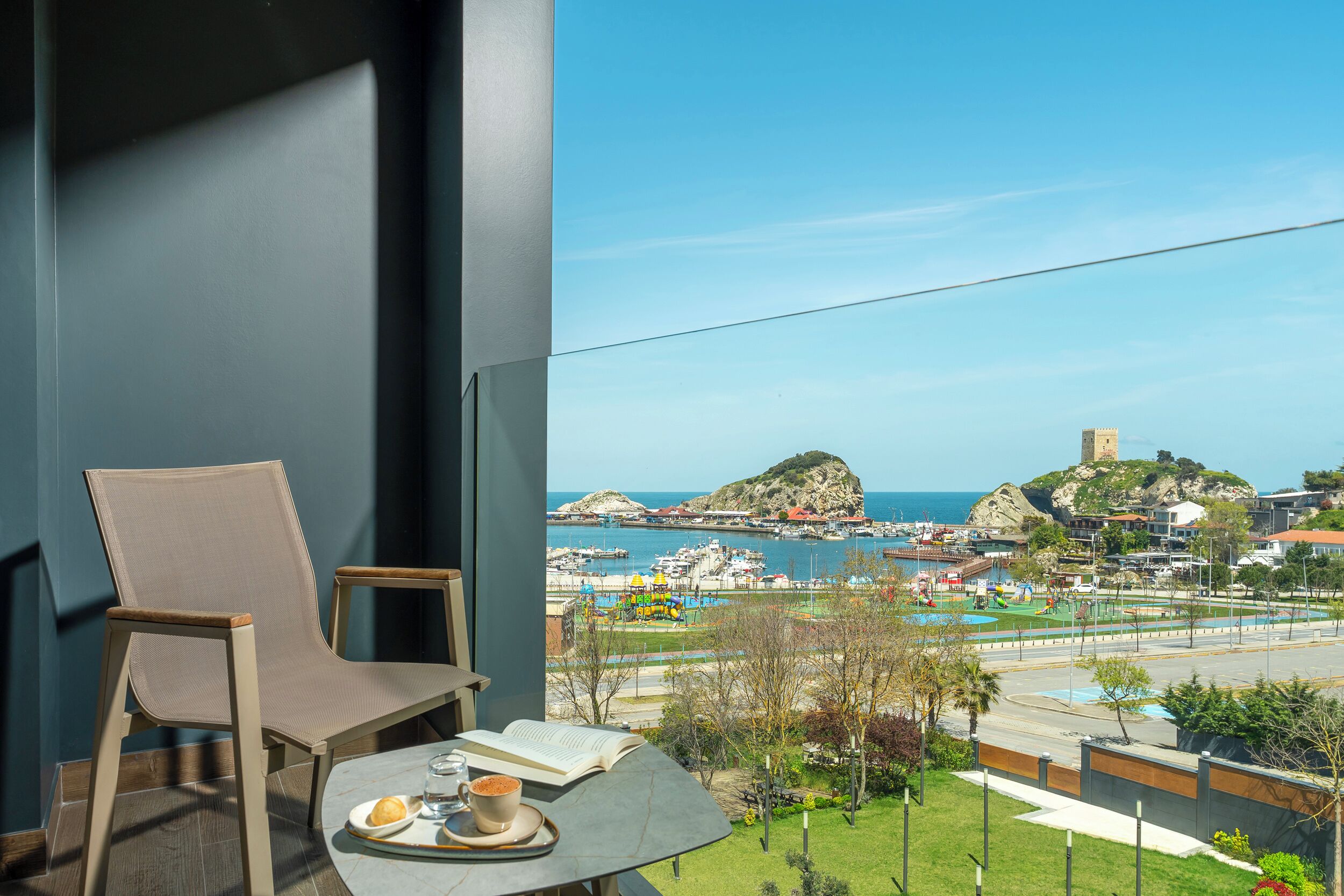 Un balcón con una silla y una mesa, con vista a un puerto deportivo y una costa rocosa.