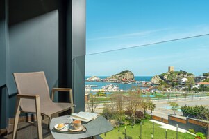Un balcón con una silla y una mesa, con vista a un puerto deportivo y una costa rocosa.