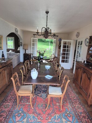 Ein Esszimmer mit einem großen Holztisch, Stühlen, einer Kandelaber und Blick auf den Garten durch geöffnete Türen.