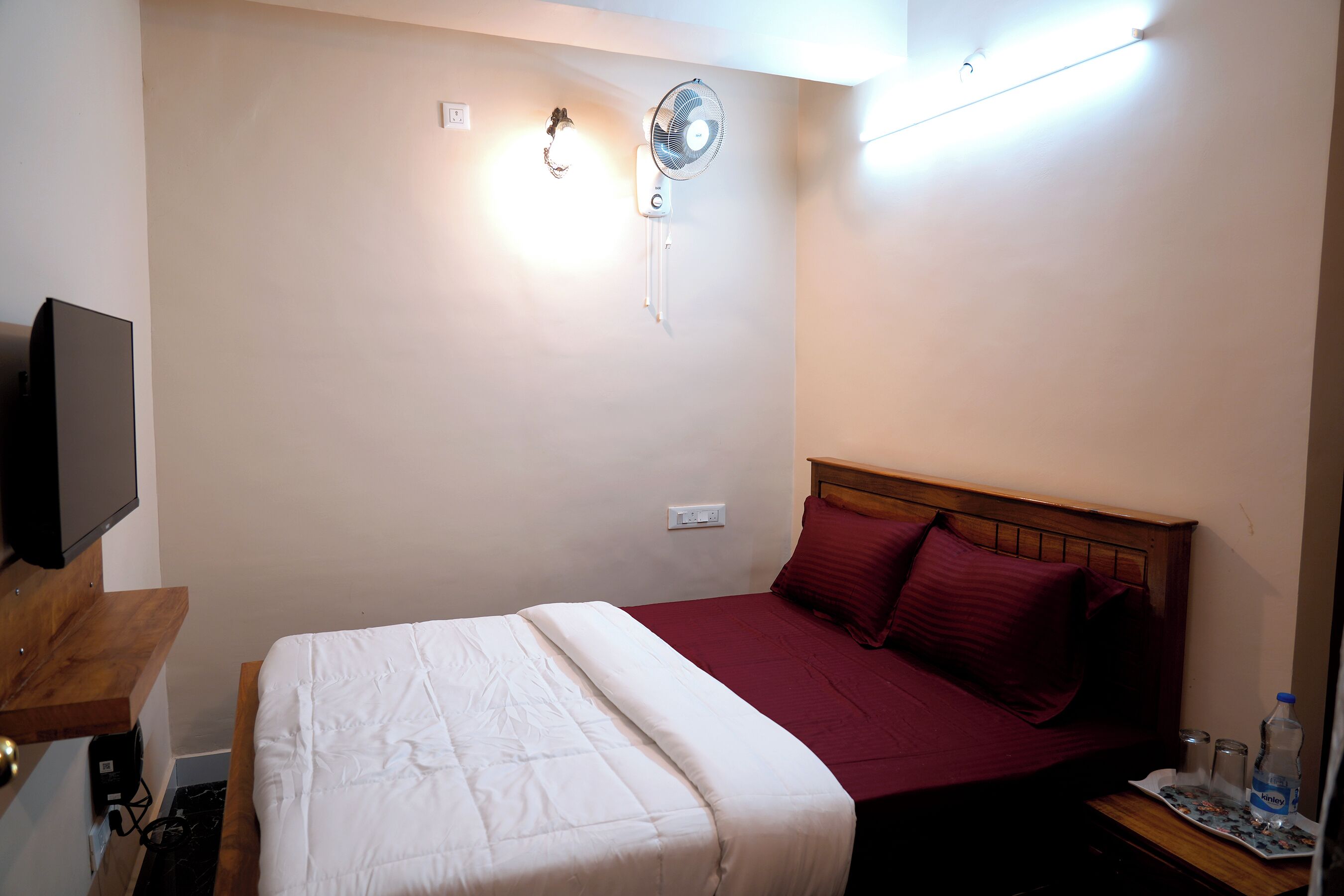 A hotel room with a bed, a bedside table with a bottle, a wall-mounted fan, and a TV on a stand.