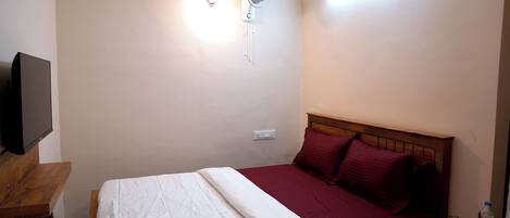 A hotel room with a bed, a bedside table with a bottle, a wall-mounted fan, and a TV on a stand.