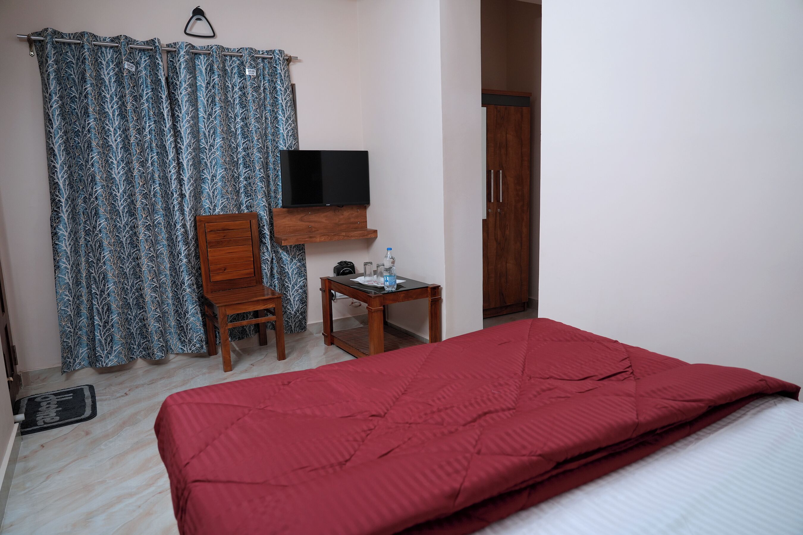 A room with a bed, a television on a wooden shelf, a small table with a bottle, and a wardrobe.