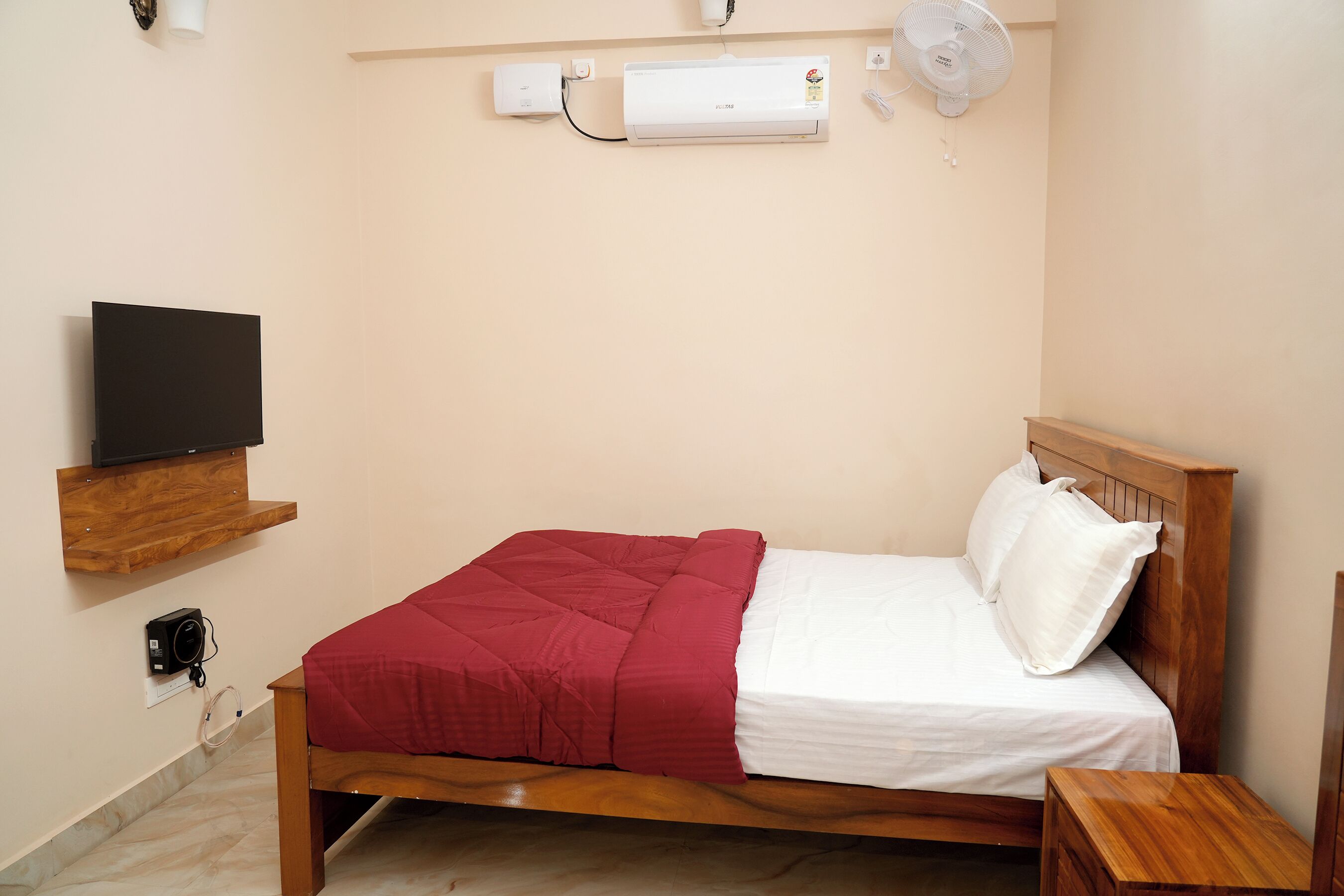A neatly made bed with a red blanket and white pillows, a wooden headboard, a bedside table, a mounted TV, and an air conditioning unit on the wall.