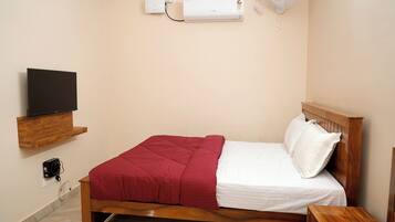 A neatly made bed with a red blanket and white pillows, a wooden headboard, a bedside table, a mounted TV, and an air conditioning unit on the wall.