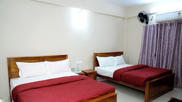 Habitación de hotel con dos camas de madera, cada una con ropa blanca de cama y mantas rojas. Hay una mesita de noche con una botella de agua y un ventilador en la pared.