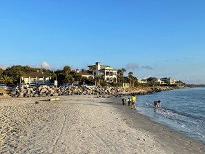 Een strand met een rotsachtige kustlijn, een pier en een rij huizen op de achtergrond.