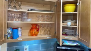 A kitchen with wooden cabinets containing wine glasses, plasticware, and storage containers. - Easy Beach Stay For Families + King Bed + Bikes + Pool! (St. Simons Island)