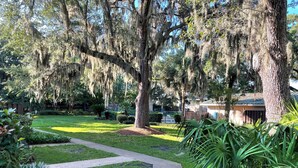 A well-maintained garden with a paved walkway, mature trees with Spanish moss, and a small building with a sloped roof. - Easy Beach Stay For Families + King Bed + Bikes + Pool! (St. Simons Island)