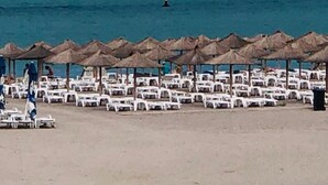 Ein Strand mit Reihen weißer Liegestühle und strohgedeckten Sonnenschirmen, einem klaren blauen Meer und einem Schiff in der Ferne.