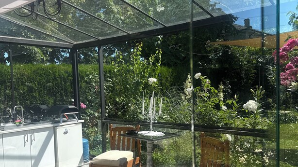 Eine verglaste Terrasse mit Küchenzeile, Sitzgelegenheiten im Freien und Blick auf einen üppigen Garten.