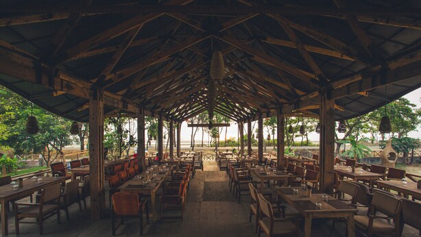 An open-air restaurant with wooden tables and chairs, a high ceiling with a geometric pattern, and a view of the outdoors. - Anchor Eco Resort & Spa (Cox's Bazar)