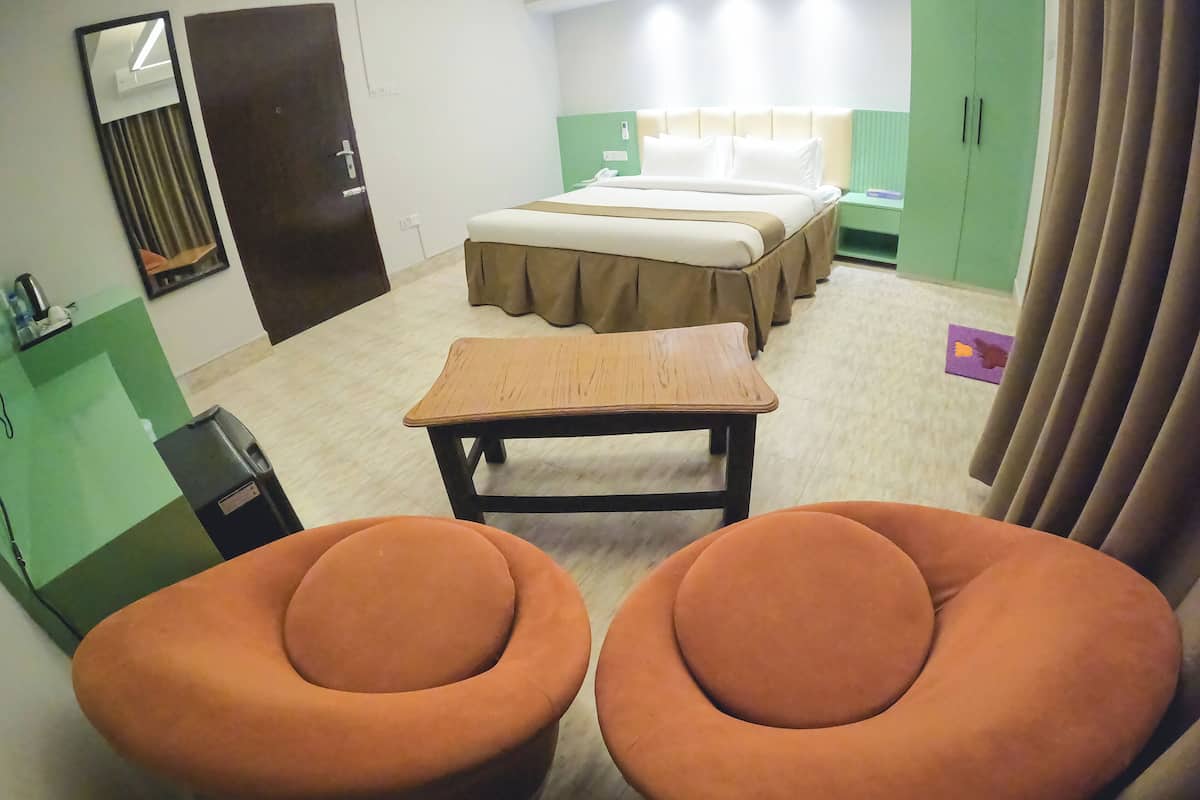 A hotel room with a bed, a desk, two orange chairs, a green cabinet, and a mirror.