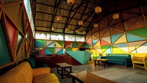 A room with a high ceiling, wooden beams, and colorful geometric wall panels. There are sofas, a table, and hanging baskets. - Anchor Eco Resort & Spa (Cox's Bazar)