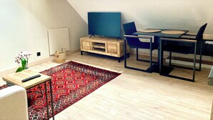 Ein modernes Loft-Zimmer mit einem Holzesstisch, blauen Stühlen, einem Fernseher auf einem Holzschrank, einem roten Teppich mit Muster und einem kleinen Beistelltisch mit Blumen in einer Vase.
